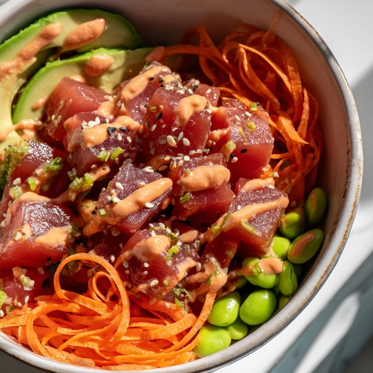 A close-up of a Spicy Tuna Poke Bowl with Sriracha Mayo features marinated raw tuna cubes, sliced avocado, and crisp vegetables atop fluffy rice, drizzled with creamy pink sauce.