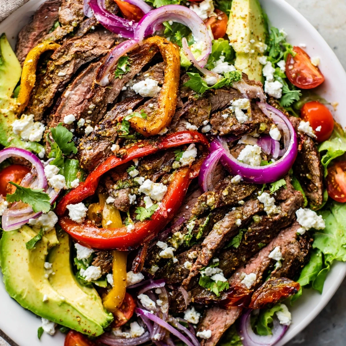 Close-up of juicy Beef Fajita Salad topped with avocado, crumbled cotija cheese, and cherry tomatoes on a white plate.
