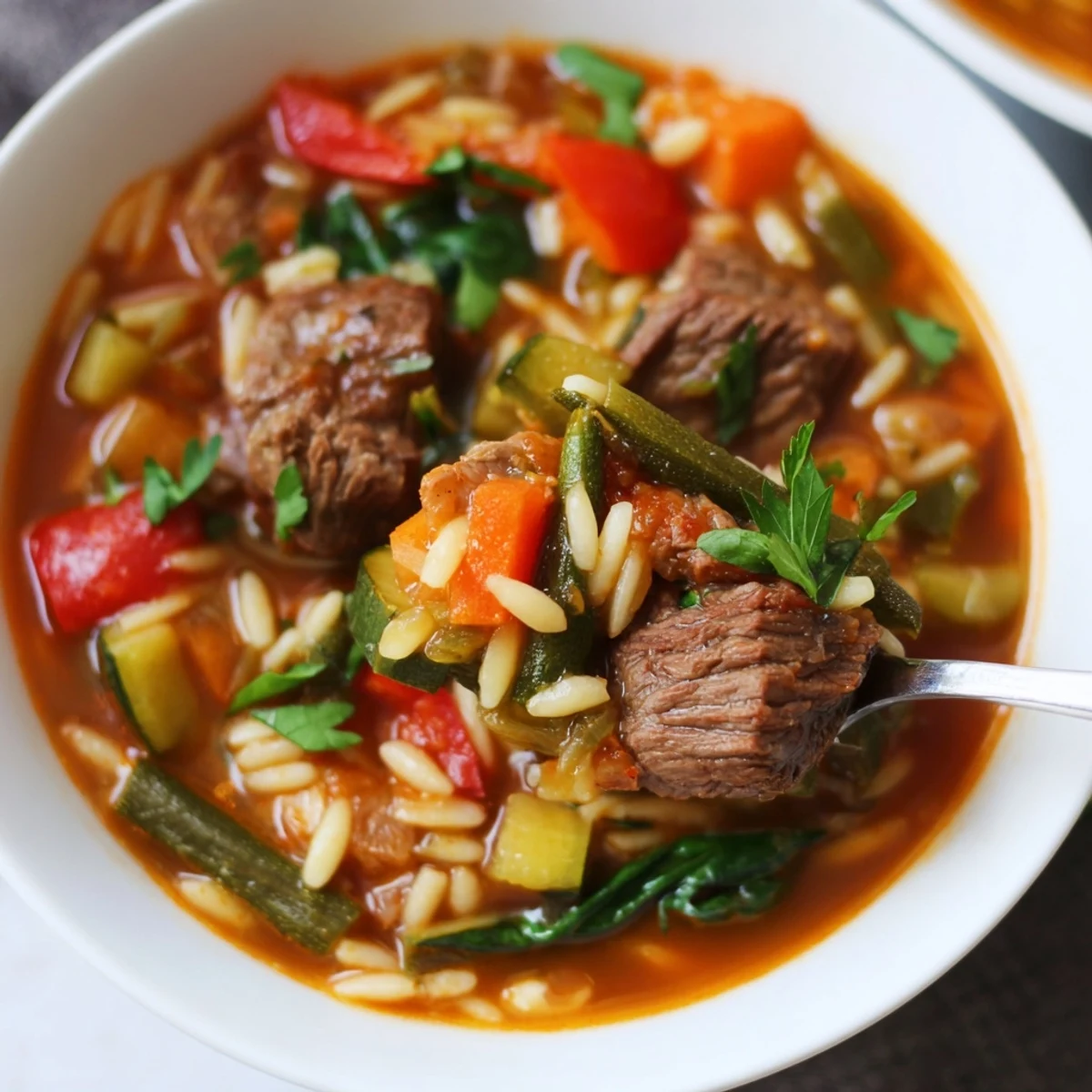 Spoon lifting tender beef and orzo from rich vegetable soup in ceramic bowl