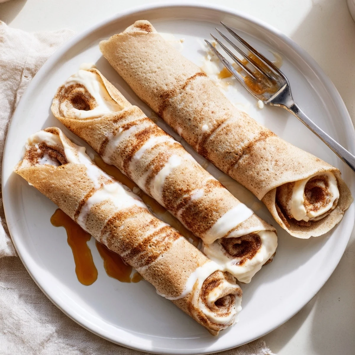 Cinnamon Roll Protein Crepes drizzled with cream cheese glaze on a warm plate