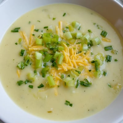 A close-up of smooth Irish Potato Leek Soup with Cheddar, garnished with grated cheese and chives, steaming in a white ceramic bowl.  